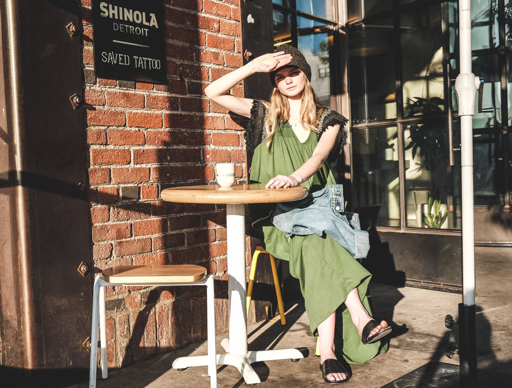 A woman sitting at a small outdoor table covering her eyes from the sun. She's wearing our Zoe Dress in Cactus and a dark grey Uplifters beanie. She also has a jean jacket laid across her crossed legs.