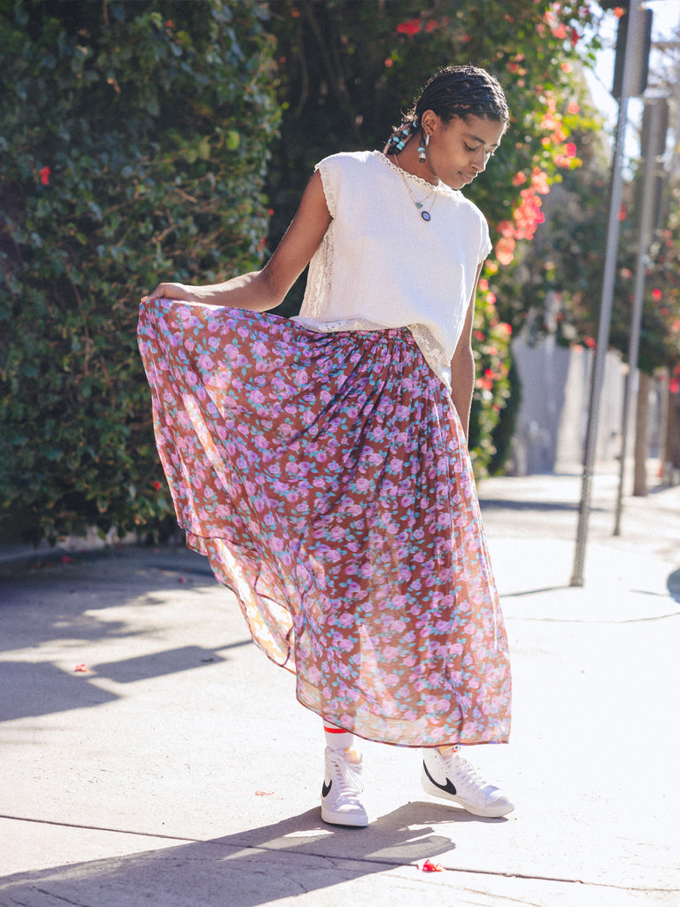 Woman is standing looking at her Nike shoes while holding one side of her Penny midi skirt in Rose Cafe, wearing the Mira blouse. She is standing on the side walk in front of a large hedge.