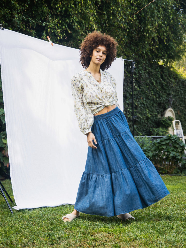 A women Afro standing facing the camera with her hip popped the right with the wind blowing her skirt out. She is wearing the Frida skirt, a blouse tied up at her stomach, and sandals. She is standing against a white sheet backdrop outside with grass and trees behind her