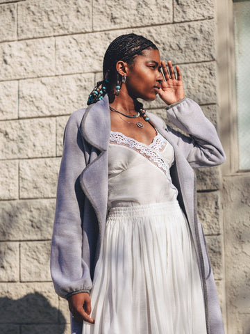 Waist up of a woman wearing our Jane Duster Coat in Heather Grey Fleece with a darker grey trim, the white Rio cami, and white Penny Maxi Skirt. She's looking to the side due to the sun in front of a light beige stone wall.