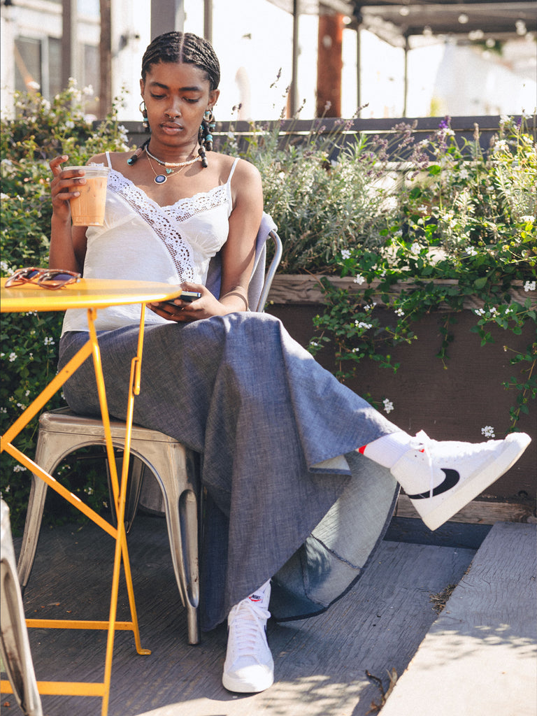 A dark complexion woman with braids sitting at a small yellow outdoor table with her legs crossed a to-go iced latte in one hand and her phone in the other. She is wearing a white cami with lace, the Joanie bias cut skirt and high top white Nikes. Behind her is planters full of green plants and small flowers, it’s also very sunny outside