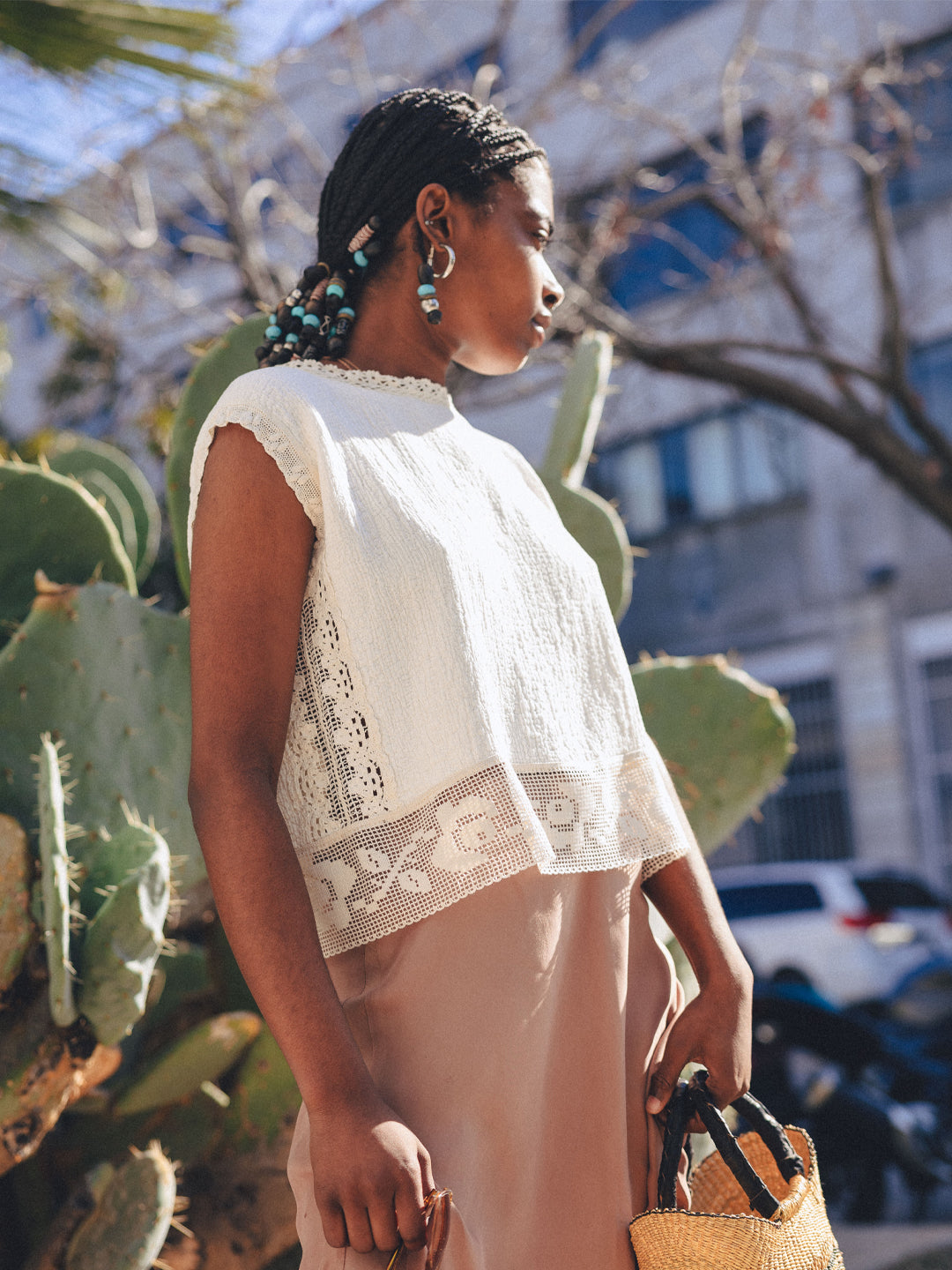 Woman standing in front of a large cactus in a cream boxy short sleeve top with vintage lace trim at the bottom, down the sides, the neck and arms. She is also wearing a light pink skirt and holding a basket purse.