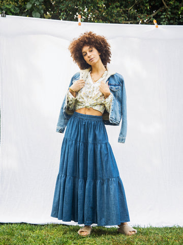 A women with a small Afro standing facing the camera with her head tilted and hip popped the the left. She is wearing the Frida skirt, a blouse tied up at her stomach, jean jacket and sandals. She is against a white sheet backdrop outside with grass and trees behind her
