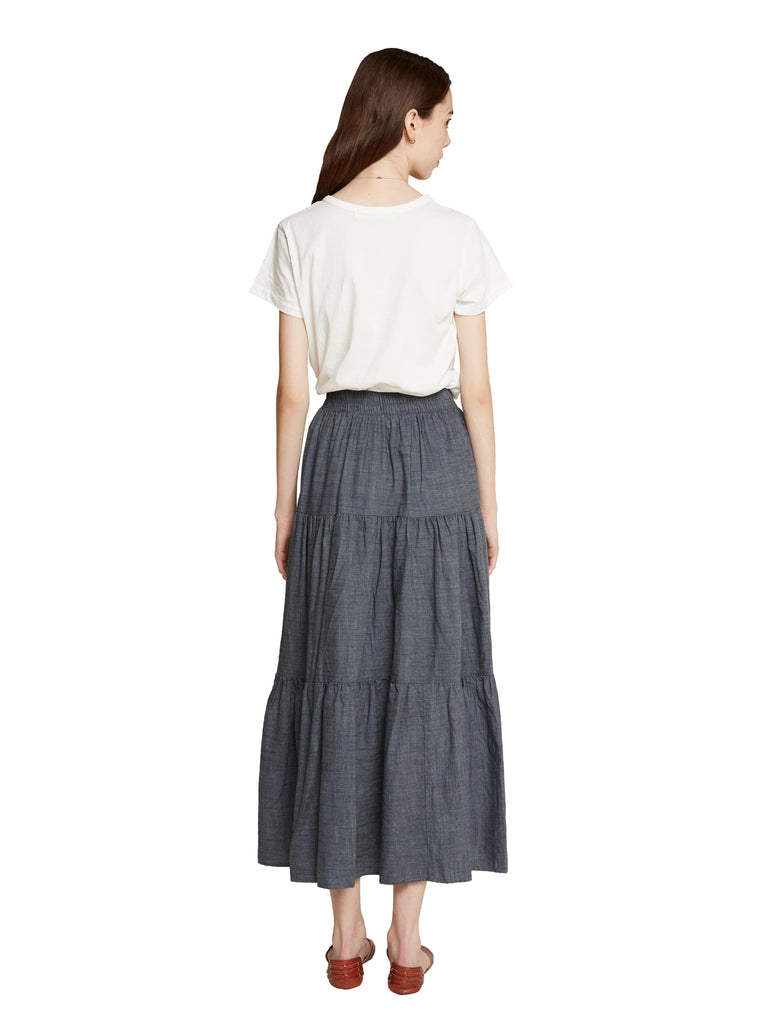 A woman standing with her back to the camera with her arms by her sides and her hair over one shoulder wearing a white tee shirt and the Frida skirt in a dark chambray color against a white background.