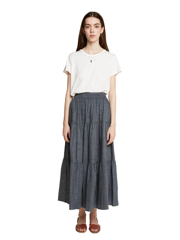 A woman standing facing the camera with her arms by her sides wearing a white tee shirt and the Frida skirt in a dark chambray color against a white background.