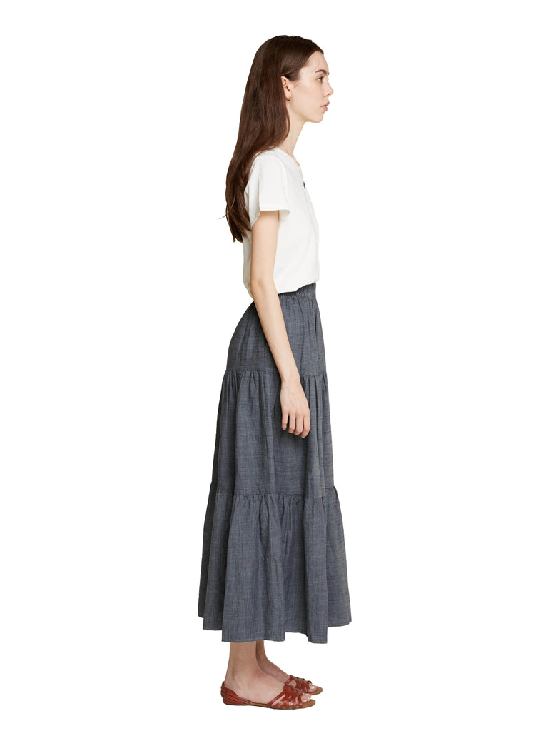 A woman standing facing the right of the camera with her arms by her sides wearing a white tee shirt and the Frida skirt in a dark chambray color against a white background.