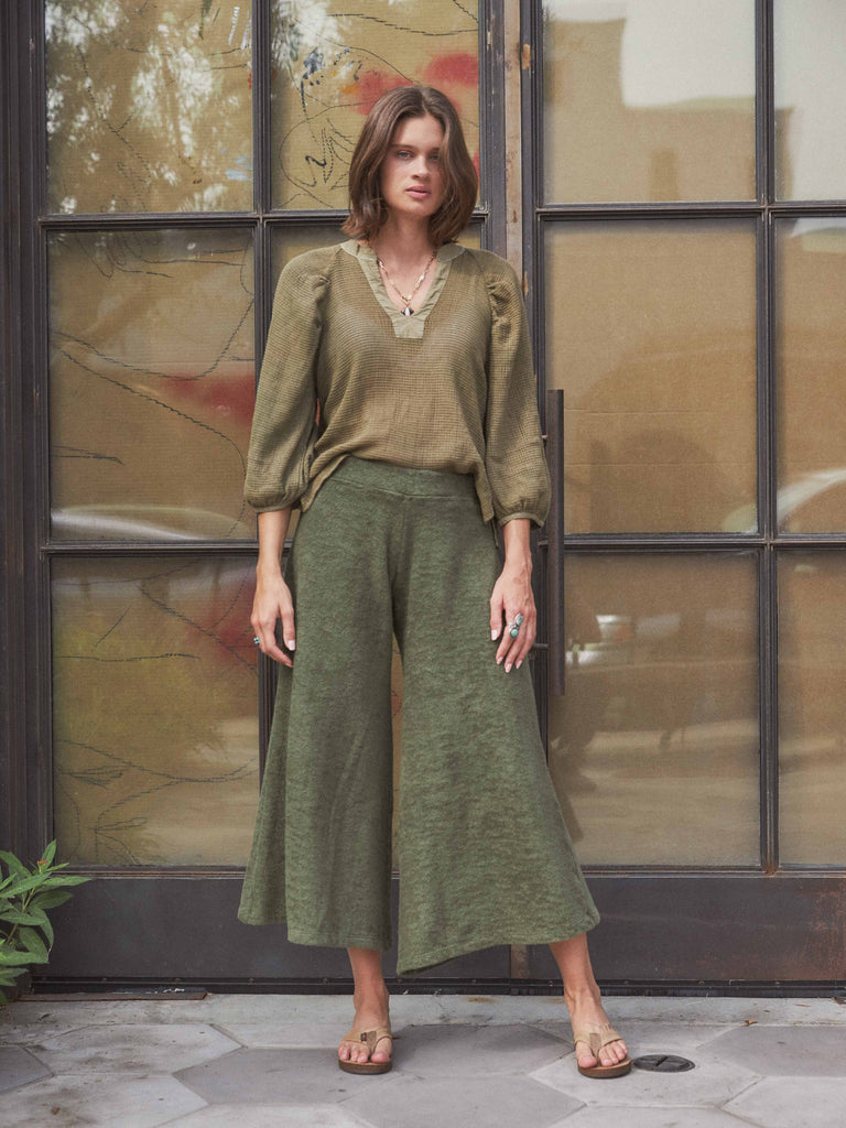 A same shot of a tanned woman but full body. She is standing with the weight on one foot and one leg out to the side with one hand by her side and the other rested on her leg. She is wearing the Paloma Pant with an army green blouse, tan sandals and two silver rings with turquoise on them. She is standing outside in front of two doors with top to bottom windows, they are covered with Kraft paper from the inside.