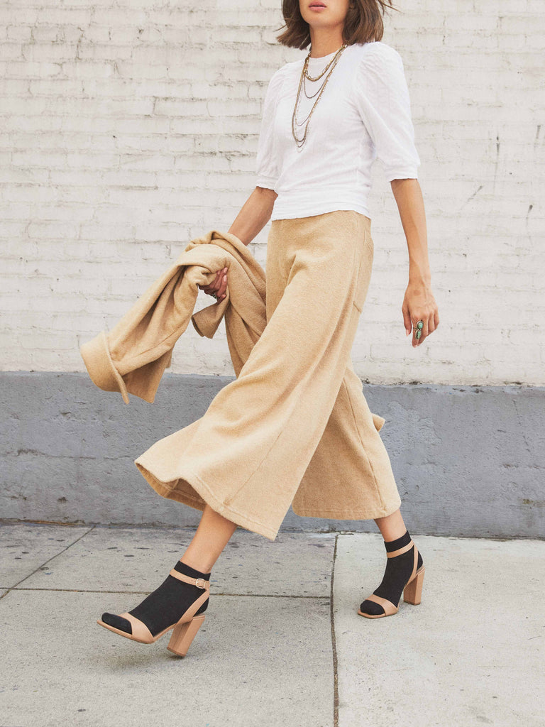 A woman is walking down the sidewalk with a coat in one hand, a white half sleeve top, wide leg Paloma Pant, tan straps heels and black socks, and long necklaces. Her face is half cut off in the photo but you can see she has short brown hair